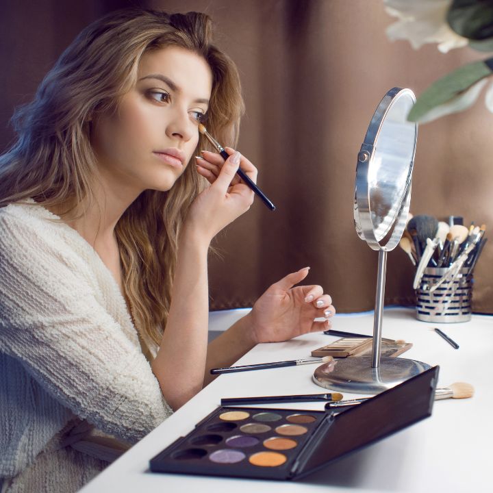 A makeup student uses her Makeup Artistry Kit to apply eyeshadow in front of a vanity mirror, with professional brushes, palettes, and cosmetics neatly arranged on the table.
