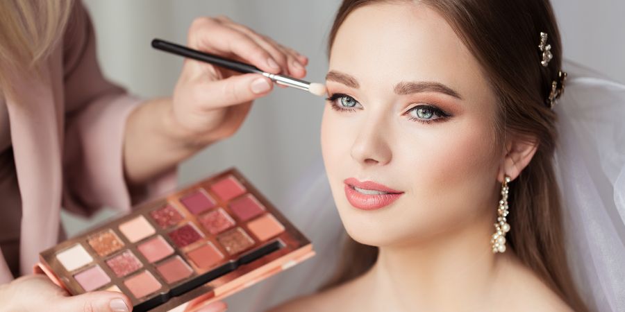 A makeup artist uses a rose-toned eyeshadow palette from their Makeup Artistry Kit to create a soft bridal look on a smiling bride.