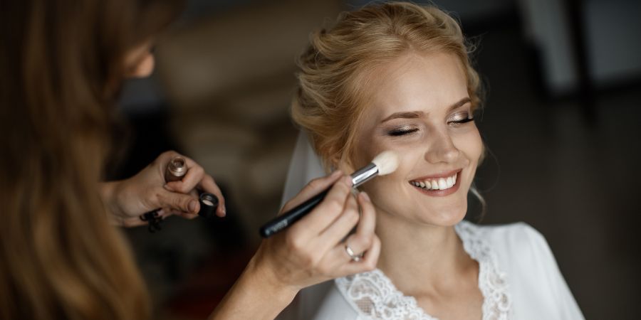Bride smiling while a makeup artist applies blush, showing the real-life pros and cons of working as a makeup artist.