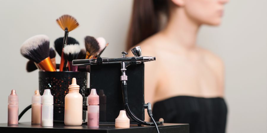 Airbrush makeup machine, foundation bottles, and professional brushes set up beside a model; visually representing tools used to explore the pros and cons of airbrush makeup in beauty applications.