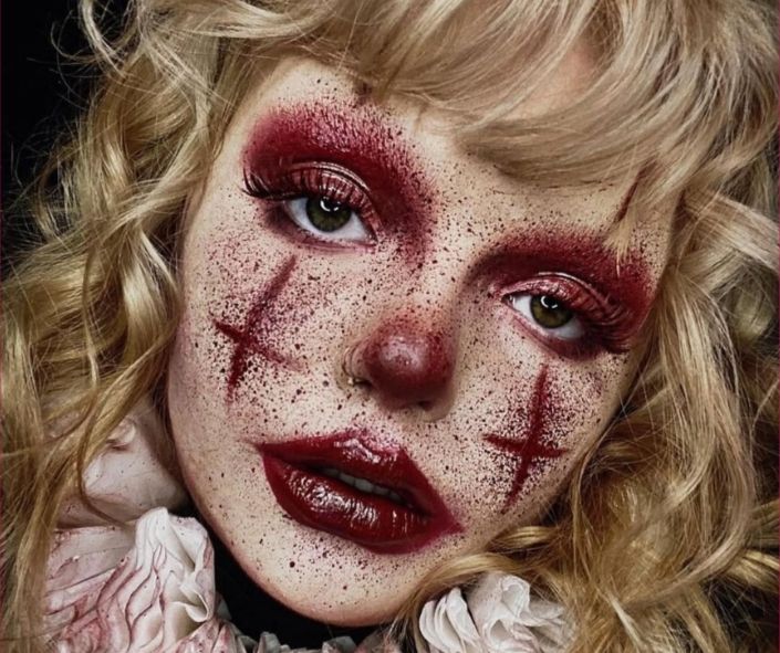 Scary Halloween makeup featuring a creepy clown look with red smeared eyeshadow, bold cross designs, dark red lips, and dramatic curls for a haunting effect.