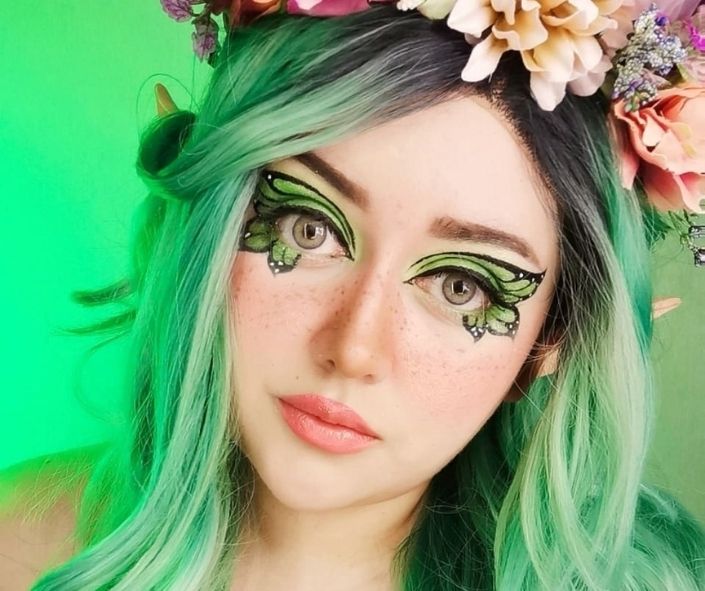 Whimsical Halloween makeup idea featuring green butterfly eye makeup, pastel green hair, and a floral crown for a magical fairy-inspired look.