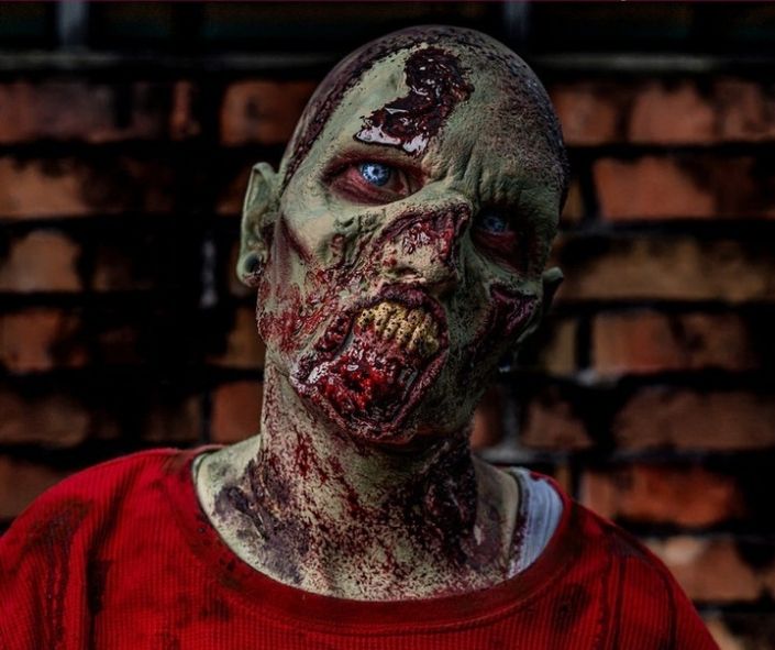 Scary Halloween makeup featuring a gruesome zombie look with rotting green skin, fake blood, decayed teeth, and eerie blue contact lenses for a terrifying effect.