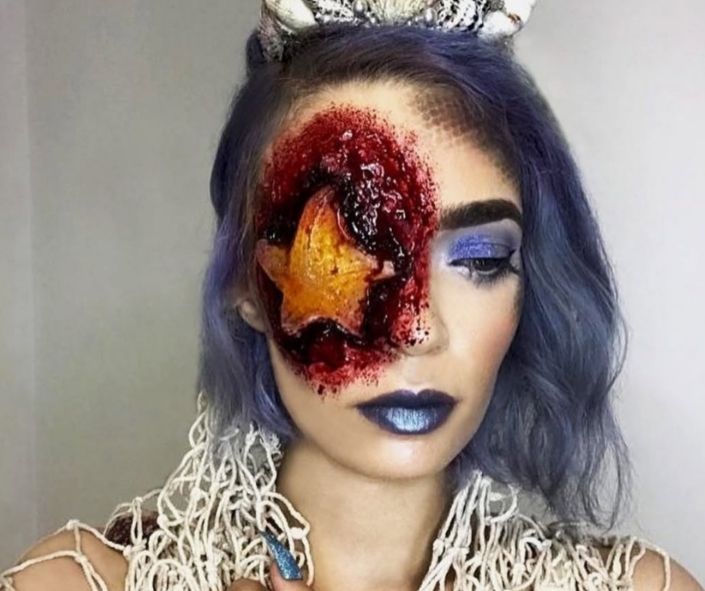 Scary Halloween makeup featuring a gruesome starfish prosthetic over one eye with bloody special effects, paired with metallic blue lipstick and bold purple eyeshadow for a creepy ocean-inspired look.