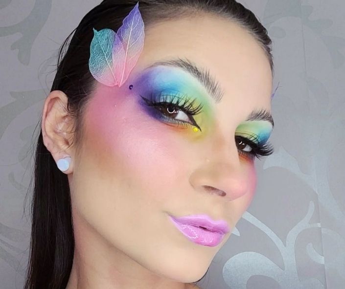 Colorful Halloween eye makeup look featuring rainbow eyeshadow, bold lashes, glossy pink lips, and delicate leaf accents for a creative costume idea.