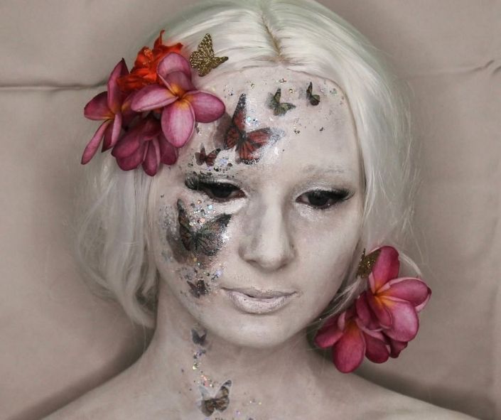 Unique Halloween makeup idea featuring a white-painted face adorned with butterfly designs, glitter accents, and bright tropical flowers for a striking fantasy-inspired look.