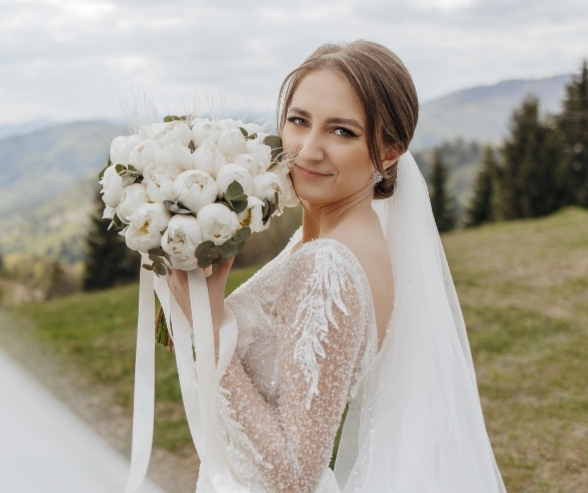 Smiling bride holding a white bouquet in a mountain setting, wearing soft glam dramatic bridal makeup with defined eyes and nude lips.