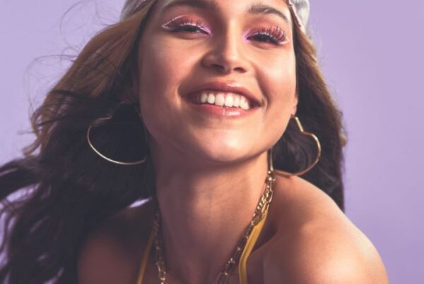 Smiling woman wearing pink shimmer eyeshadow, glossy lips, and gold jewelry, showcasing a fun and youthful look for makeup inspiration and creative artistry.