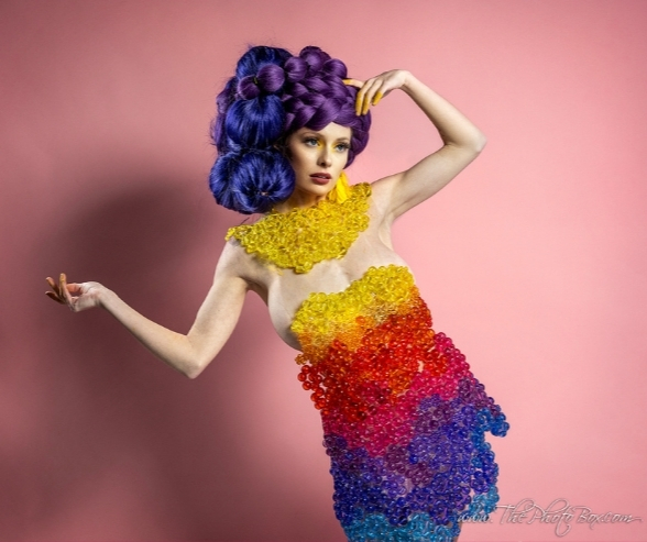 Model posing in a rainbow beaded dress and bold purple hair against a pink background, showcasing vibrant artistic makeup inspiration and creative fashion design. Makeup by QC Makeup Academy student, Kate Loveland.