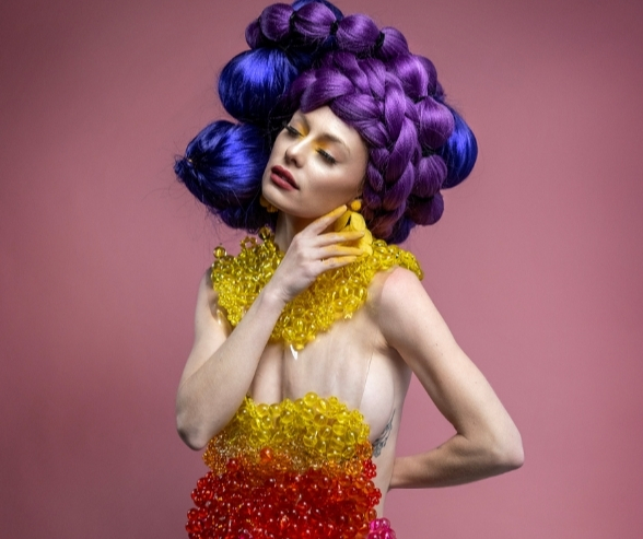 Close-up of a model wearing bold artistic makeup, yellow and red beaded fashion, and a sculpted purple hairstyle, representing vibrant makeup inspiration for creative artists. Makeup by QC Makeup Academy student, Kate Loveland.