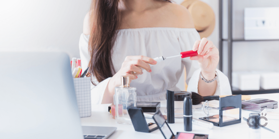 woman opening tube of makeup, behind table with laptop and other cosmetic products