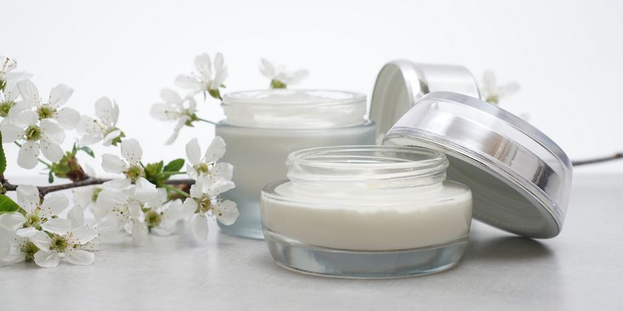 Beauty cream in a glass jars on a light gray background. Decorated with white spring flowers. Unbranded skincare product. Cosmetic cream. Close up, selective focus, side view.