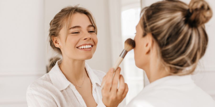 Happy fair-skinned young lady applying makeup with brush to woman in white shirt indoors. Blonde girls enjoy girls day. Cosmetic products, concept. Makeup business, makeup artist working on client.