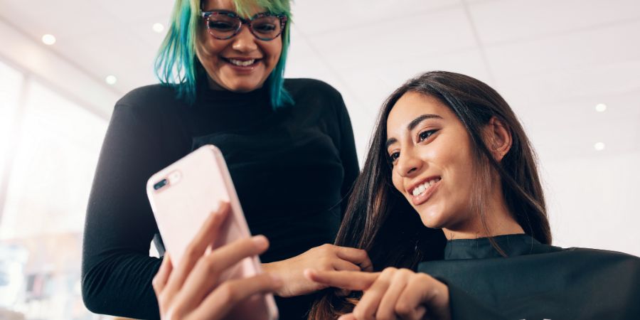 Hair stylist in colorful hairstyle looking at mobile phone while working on woman's hair. Woman showing her preferences to the hairstylist in mobile phone at the salon.