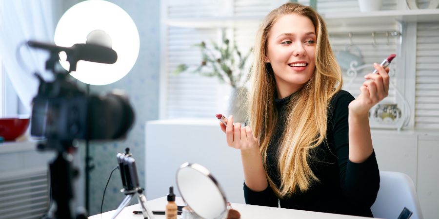 Beauty blogger woman filming daily make-up routine tutorial near camera on tripod. Influencer blonde girl live streaming cosmetics product review in home studio. Vlogger female comparing lipsticks., Viral tiktok makeup artist article.