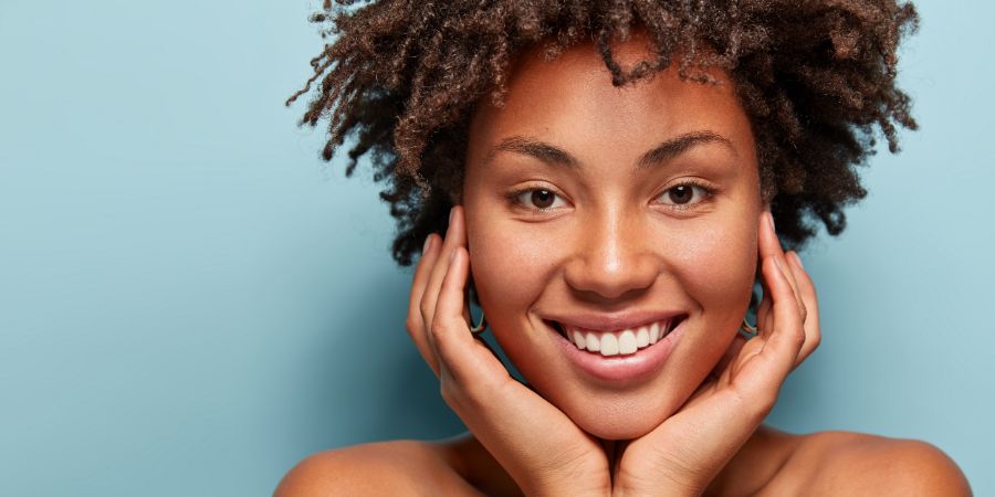 Close up portrait of relaxed black woman has gentle skin after taking shower, satisfied with new lotion, has no makeup, smiles tenderly, shows perfect teeth, stands shirtless against blue background. Beauty habits article.