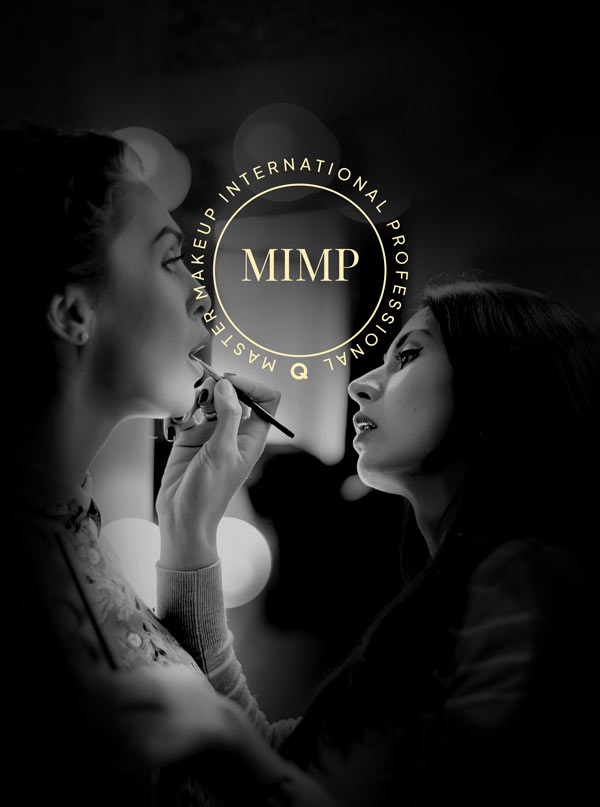 A black-and-white, vertical photograph showing a close-up of a makeup artist applying product to a model's lips. Both women are in profile, facing each other. The background is dark with soft, out-of-focus bokeh lights. Overlaid in the center is a gold circular logo featuring the acronym "MIMP" and the surrounding text "MASTER MAKEUP INTERNATIONAL PROFESSIONAL." The lighting is dramatic, highlighting the contours of their faces and creating an elegant, professional atmosphere.