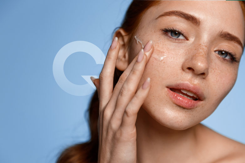 A close-up photograph of a young woman with vibrant red hair and a charming splatter of freckles across her nose and cheeks. She has luminous, dewy skin with a faint shimmer. She's applying a clear, gel-like serum to her cheek with her index and middle fingers, displaying her neatly manicured nails. The background is a clean, sky-blue color, featuring a subtly overlaid semi-transparent white capital 'Q' to the left.